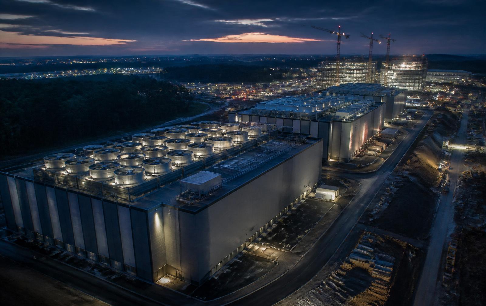 Aerial dusk view of a Northern Virginia data center corridor under construction, with illuminated cooling units and cranes on the horizon.
